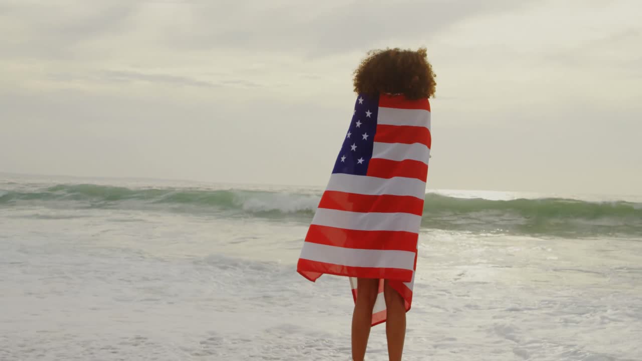 Vista posteriore di donna afroamericana avvolta nella bandiera americana sulla spiaggia 4K