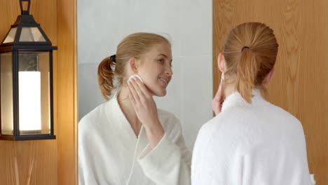 woman in bathrobe cleansing face with cotton pad while looking in mirror