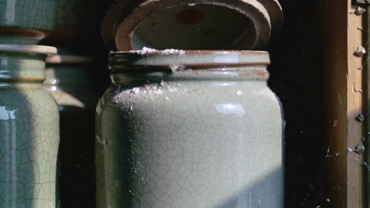 Tilting loose metal lid on green glazed jar, spraying white powder across wooden shelf and cobwebs