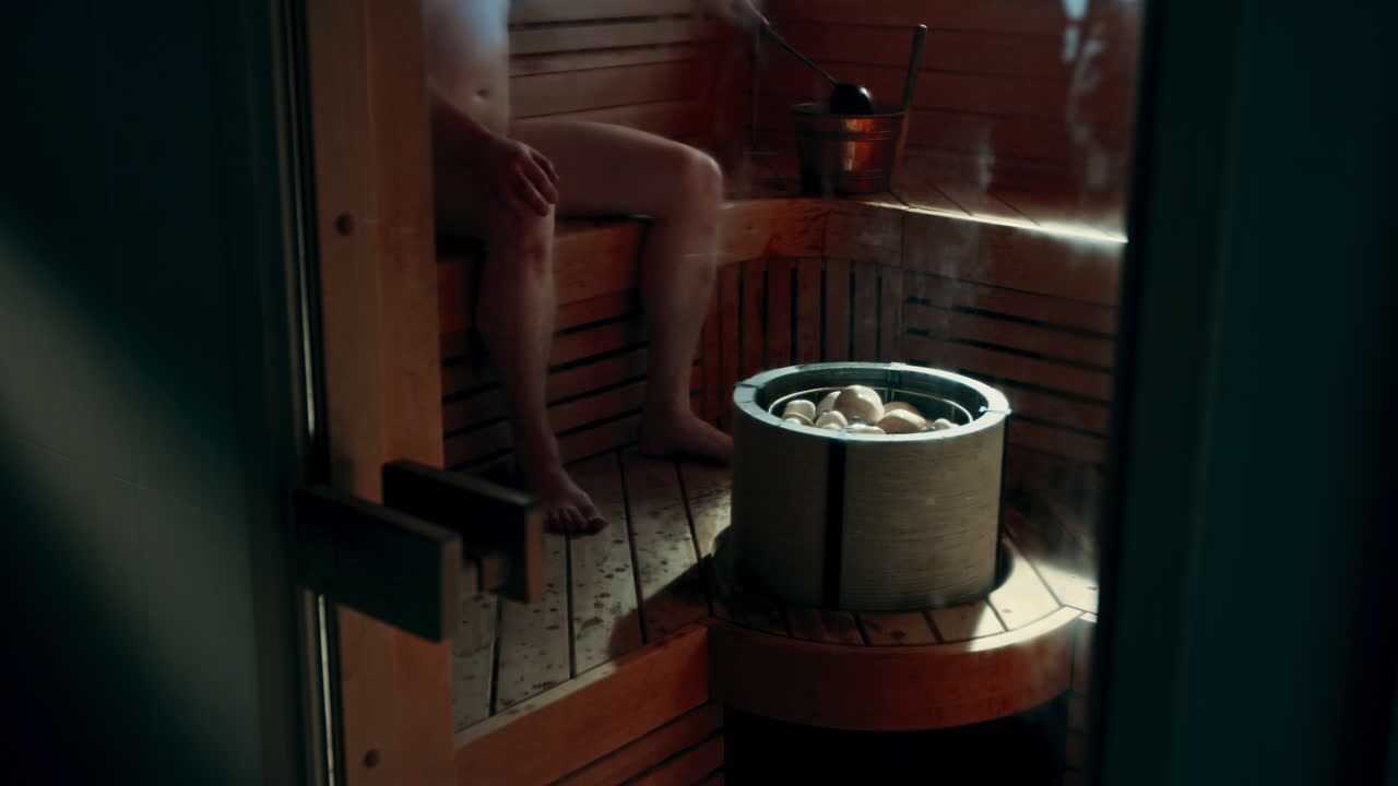 hombre sentado en la sauna, usando un cucharón o una cuchara para obtener agua del cubo de agua llamado kip o cubo, arrojando agua sobre las piedras calientes en la estufa de la sauna