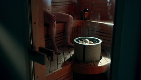 hombre sentado en la sauna, usando un cucharón o una cuchara para obtener agua del cubo de agua llamado kip o cubo, arrojando agua sobre las piedras calientes en la estufa de la sauna