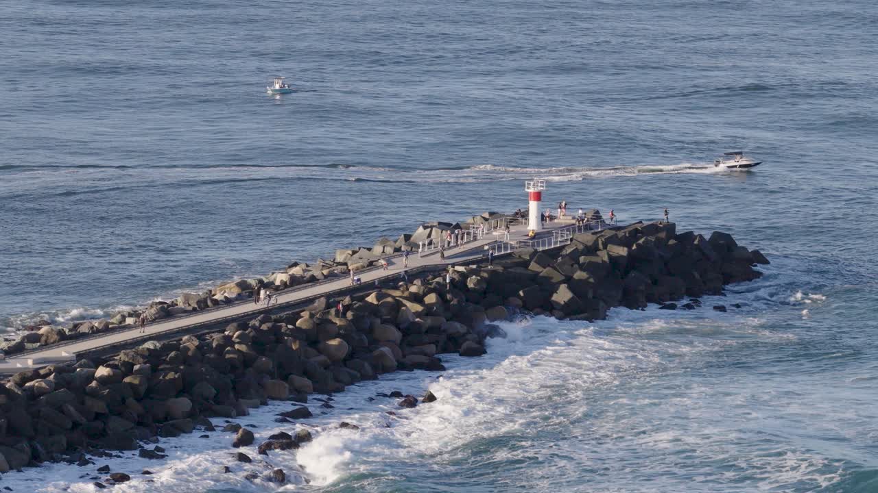 Drone footage captures boats navigating near a rocky seaway and lighthouse on the Gold Coast, Australia, under clear skies