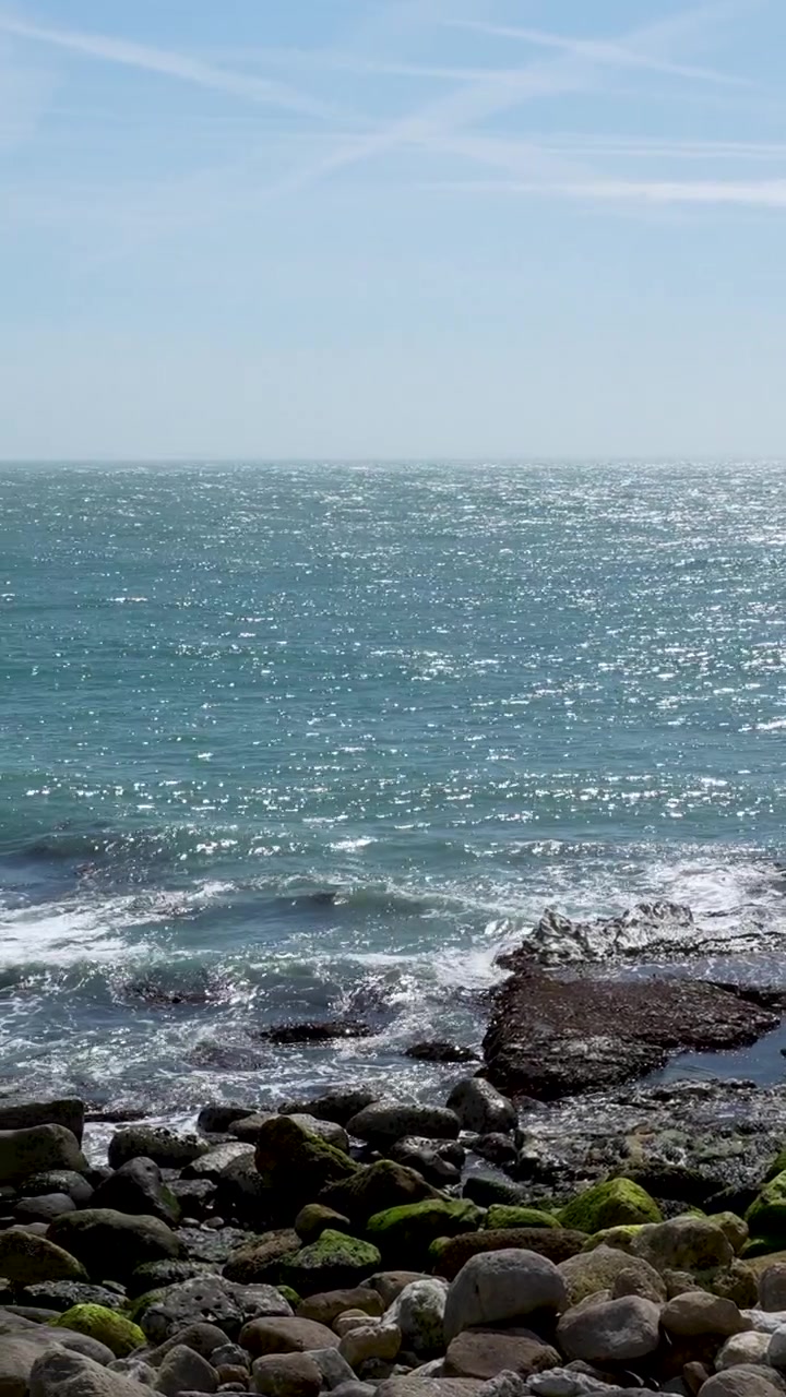 Bidikan vertikal sinematik sinar matahari terang memantul di air laut biru saat ombak menghantam garis pantai berbatu berlumut di Portland, Weymouth. Pemandangan alam pesisir yang autentik