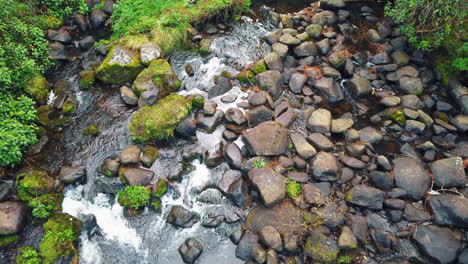 Small-water-rapids-move-gently-through-rocky-basin-below-waterfall-pond,-slowmo