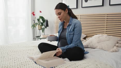 mujer caucásica en embarazo avanzado sentada en la cama, comiendo ensalada y leyendo un libro.