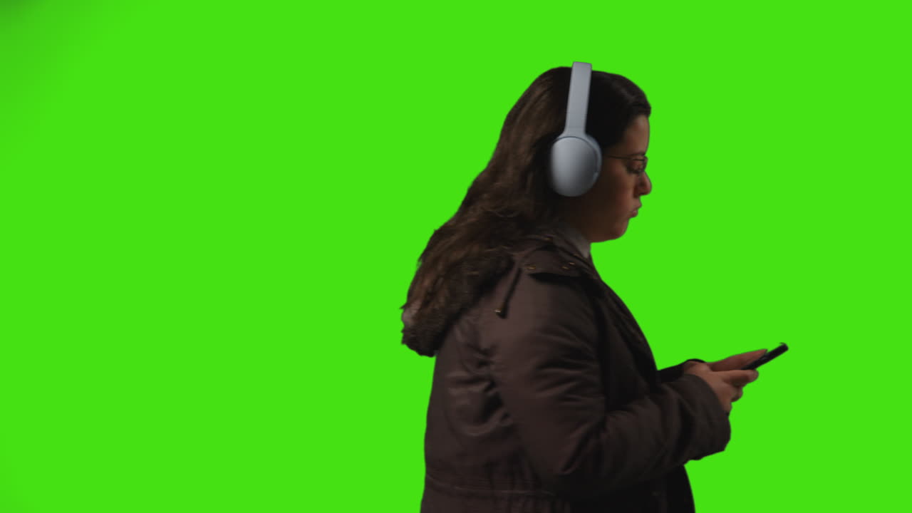 mujer joven con auriculares inalámbricos transmitiendo música desde un teléfono móvil caminando a través del cuadro contra la pantalla verde del estudio 1