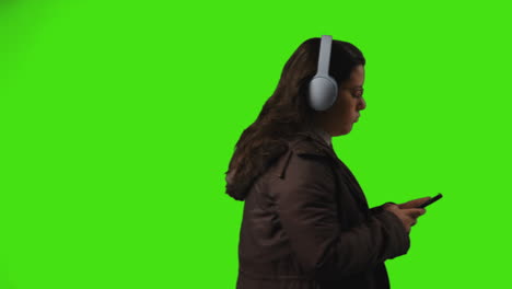 young woman wearing wireless headphones streaming music from mobile phone walking across frame against studio green screen 1