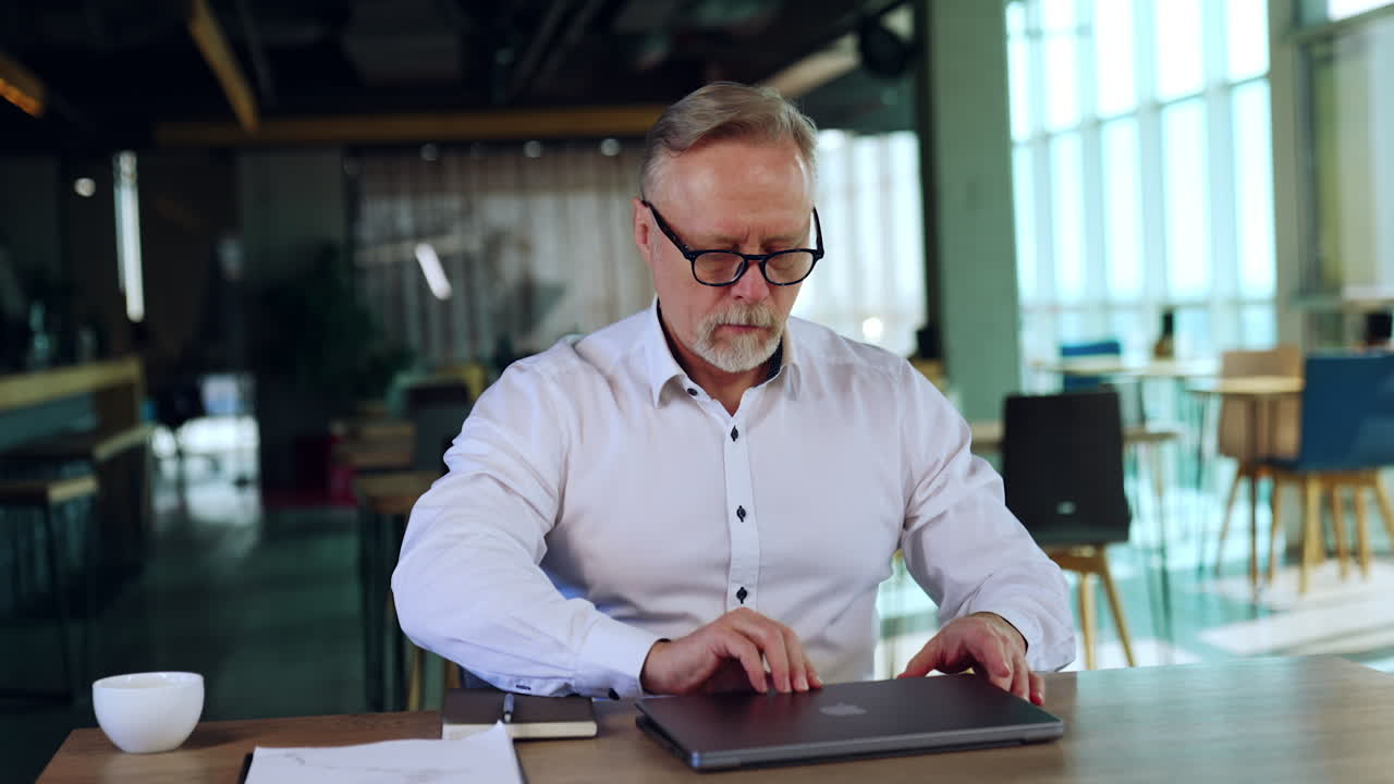 Businessman arranging his working place in the office. Man in glasses sits at opening his laptop and moving gadget and paper notebook.