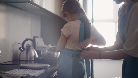 little girl is learning to cook mother is putting apron on daughter child is standing near stove