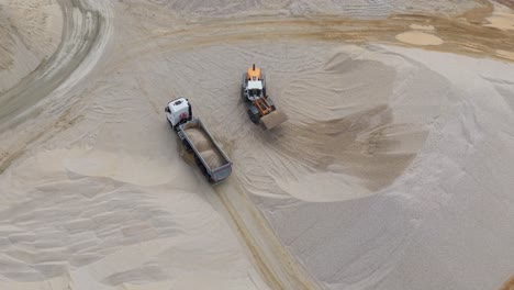 aerial view of an excavator loading gravel into a dump truck at a sand quarry. the machinery is surrounded by large mounds of sand and gravel, highlighting the scale and industrial nature of the site