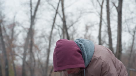 young girl in winter outfit bends slightly, feeling cold, with a blurred snowy background and bare trees, hooded jacket covers head, and chilly weather is visible in outdoor winter setting