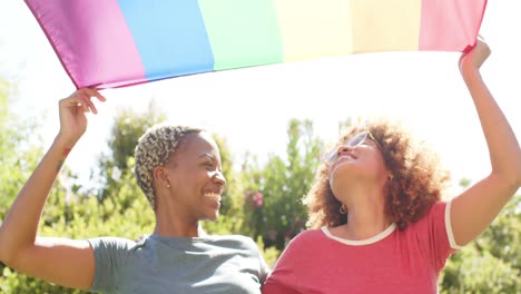 Portrait-of-happy-diverse-lesbian-female-couple-holding-rainbow-flag-of-pride-in-slow-motion