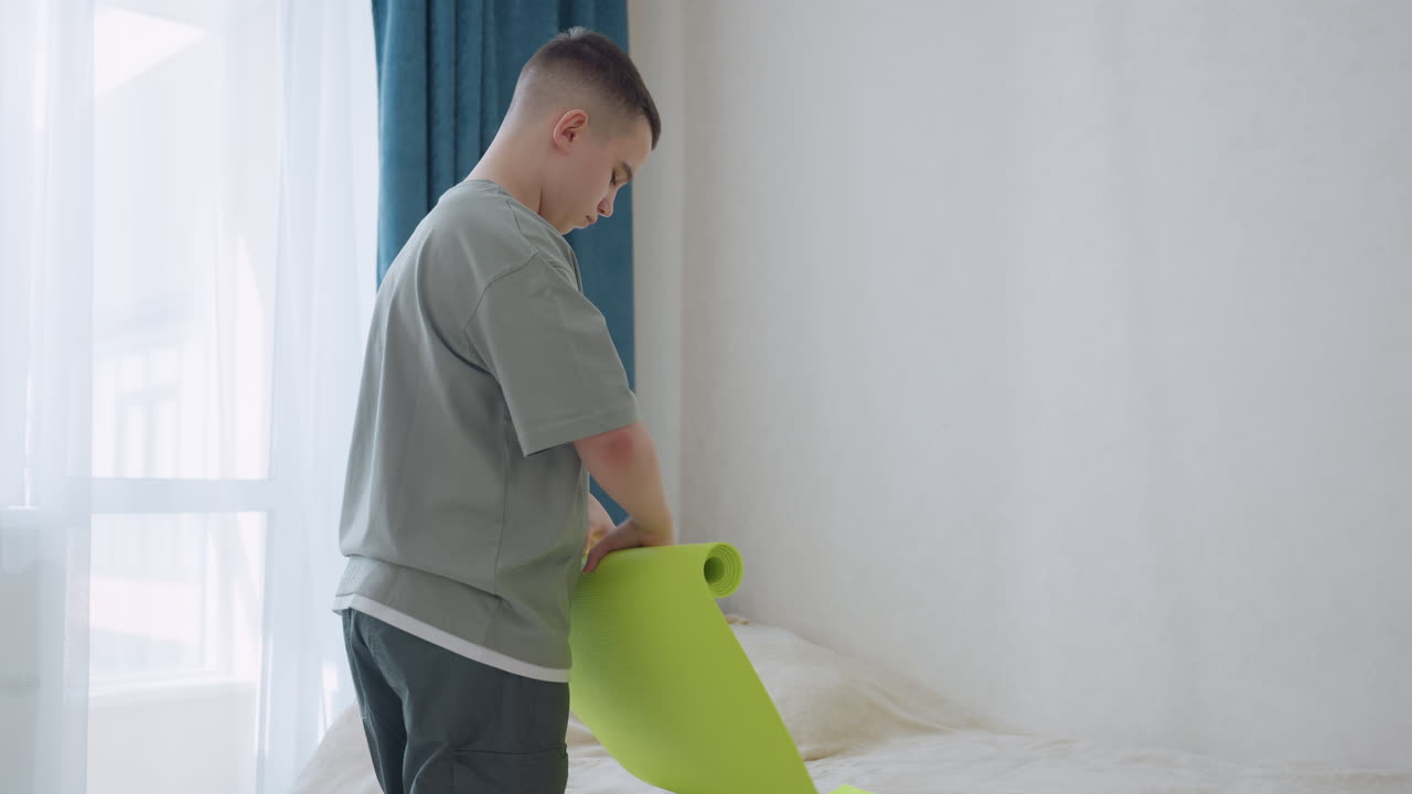 Boy in relaxed outfit folds vibrant green exercise mat with care inside sunlit room near blue curtains and bed, showing end of workout routine with quiet focus and responsibility in cozy indoor space