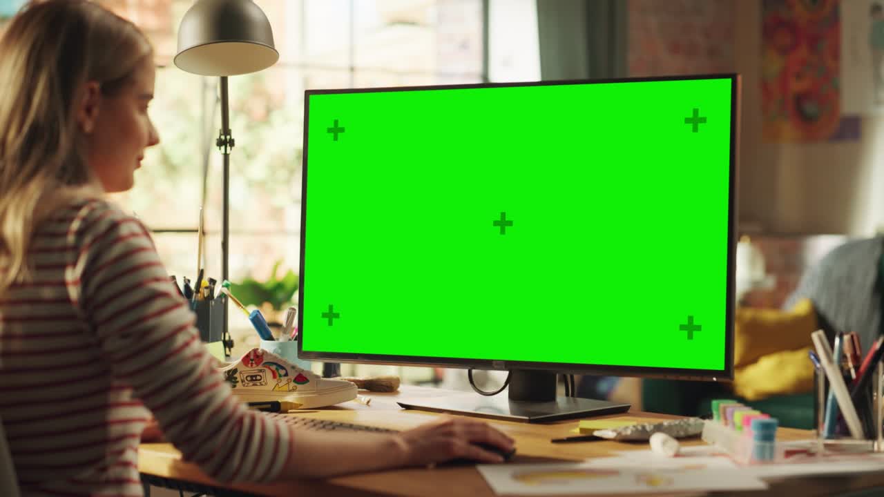 Over the Shoulder: Creative Young Woman Sitting at Her Desk Using Desktop Computer with Mock-up Green Screen. Female Caucasian Specialist Working on Computer with Chroma Key Display at Home