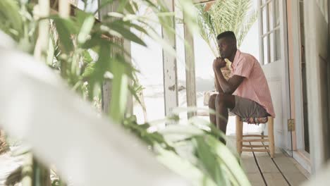 Thoughtful-african-american-man-sitting-on-porch-of-wooden-beach-house-looking-away,-slow-motion