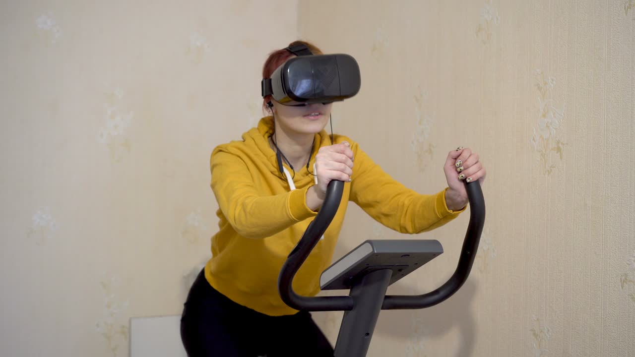Close-up of a young girl wearing virtual reality glasses doing sports at home on an exercise bike. The concept of immersion in a virtual environment. 4K.
