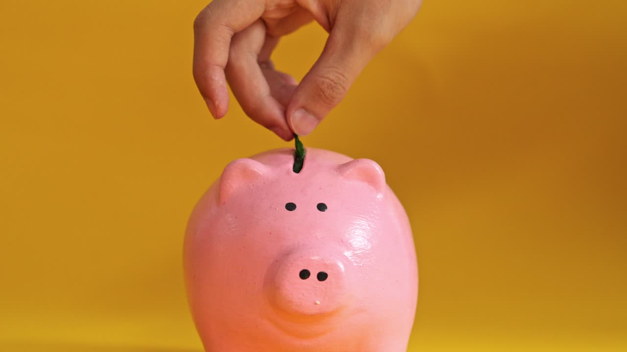 Hand putting green leaf into pink piggy bank on yellow background