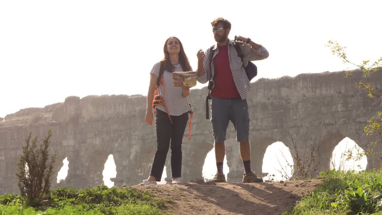 joven pareja encantadora mochileros turistas leyendo mapa señalando direcciones acueducto romano arcos en parco degli aquedotti parque ruinas en roma en romántico amanecer brumoso con guitarra y saco de dormir cámara lenta