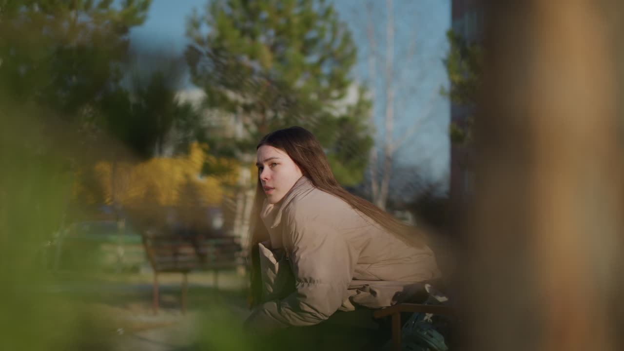 A young woman in a peach jacket is sitting alone on a park bench, with her chin resting on her hand, looking thoughtful. The background includes greenery, trees, and an urban environment