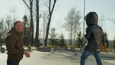 a father and son playing together outdoor as the son threw his father beanie playfully back to him, the father, dressed in a brown jacket, smiles and reaches out to catch it