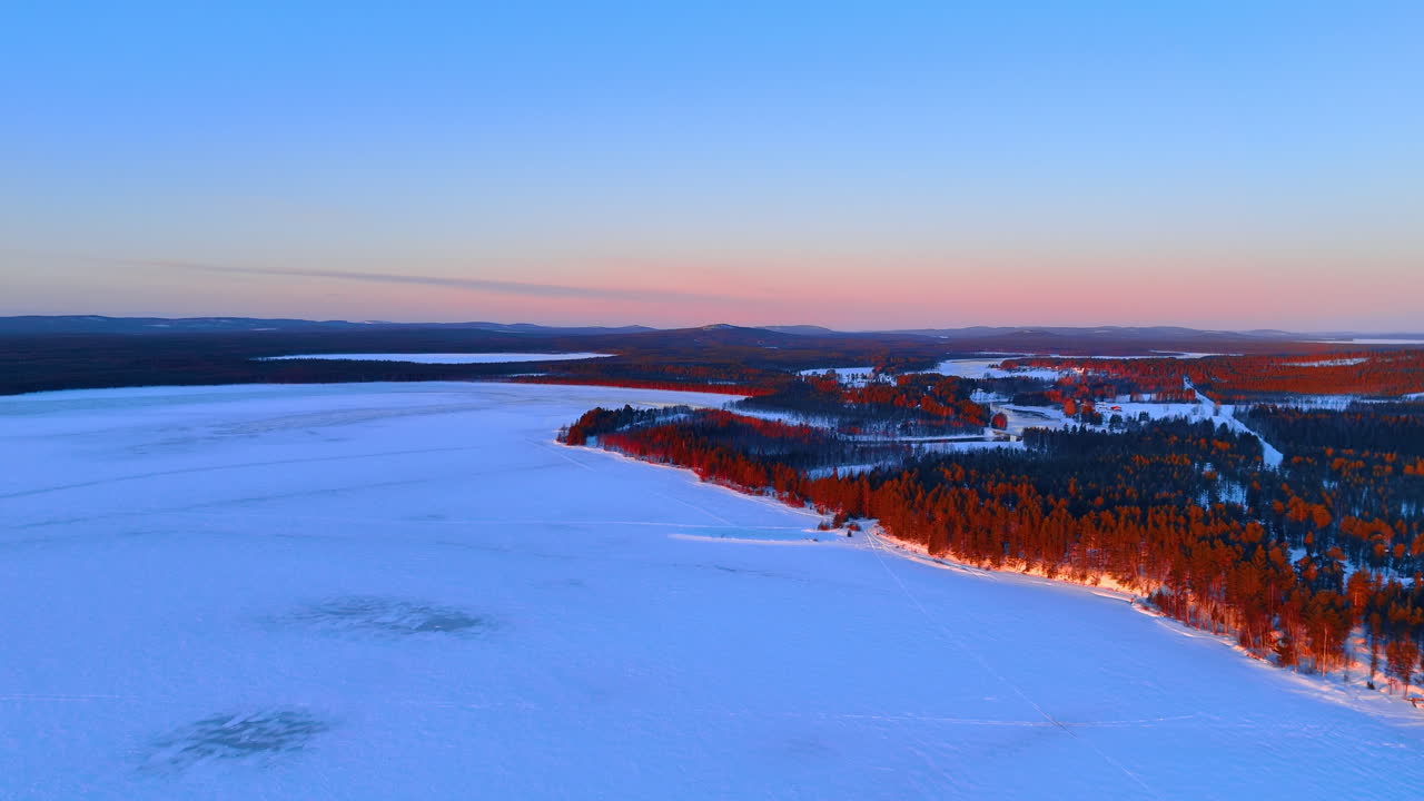 Luftfoto av snødekt skogkant langs en frossen innsjø. Oversiktsbilde av trær som kaster lange skygger på snø under en vintersolnedgang i Lappland
