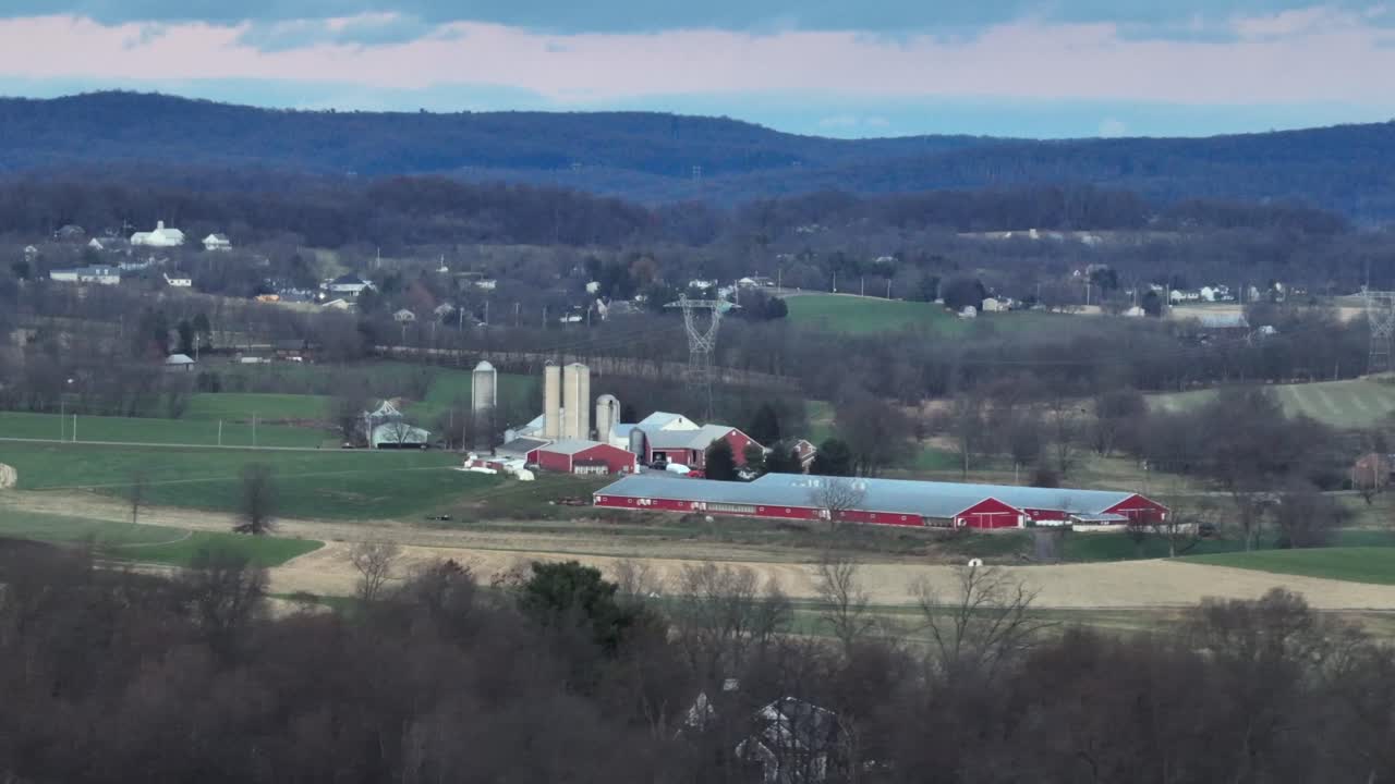 Aerial wide shot of rural American farmland showing large red barn complex with silos surrounded by rolling fields and distant hills. Scenic countryside atmosphere in quiet United States landscape