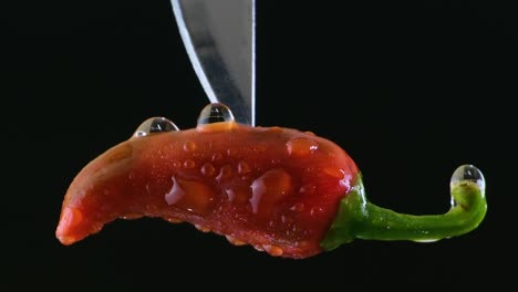 red wet jalapeno pepper pinched by a silver knife slowly dropping from top on a black background