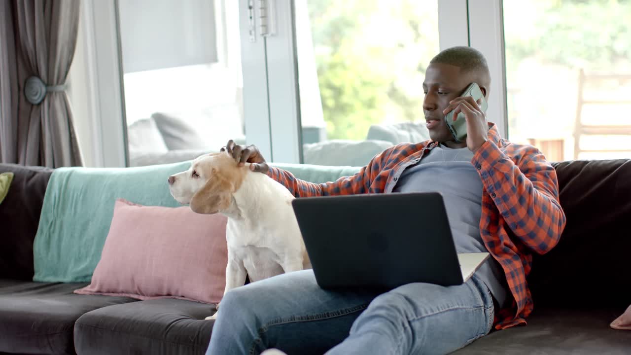Happy african american man on smartphone and using laptop at home, with his pet dog, slow motion