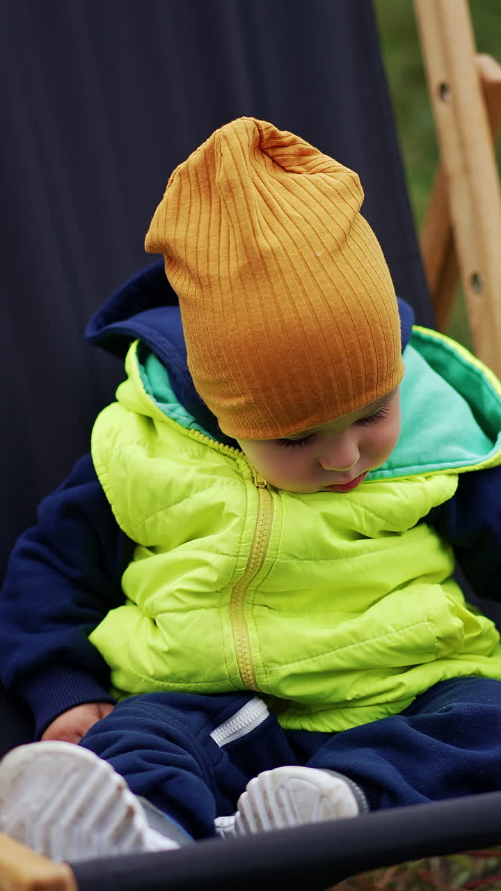 Caucasian toddler wearing warm clothes and cap sitting in garden chair. Baby looking around cautiously sitting peacefully. Close up. Blurred nature backdrop. Vertical video