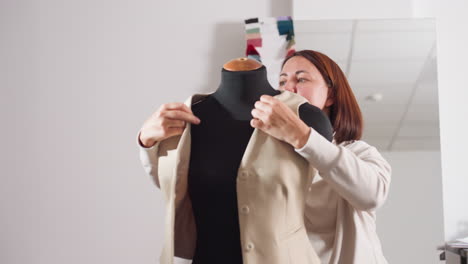 fashion designer fits buttoned beige jacket onto black lay figure, steps aside to observe result with satisfied smile, showcasing final stage of tailoring process inside bright creative workspace
