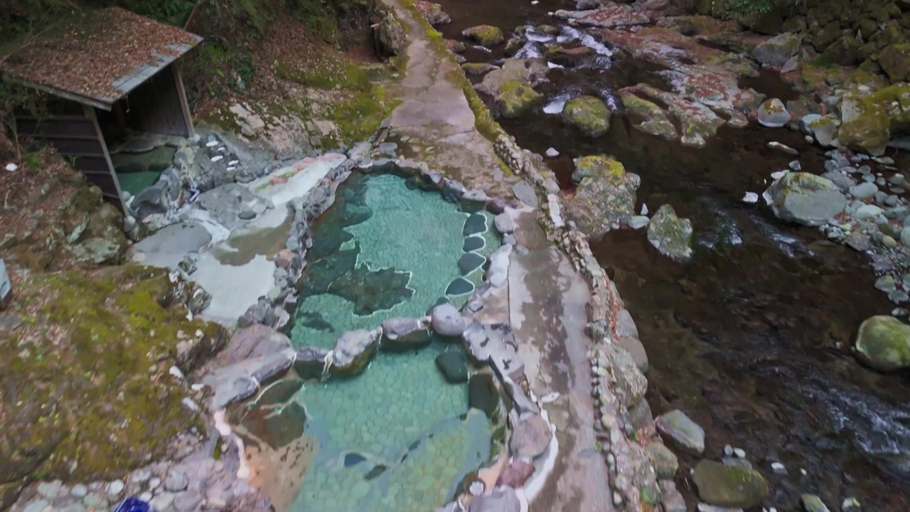 Drönarvy som flyger över traditionella utomhusbassänger med varma källor (rotenburo) med klart blått termalvatten bredvid en stenig bergsflod i Izu, Japan.