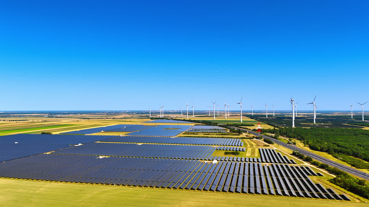 Lots of solar panels installed in the vast fields near the modern highway. Wind mills rotate in the wind at backdrop. Aerial view