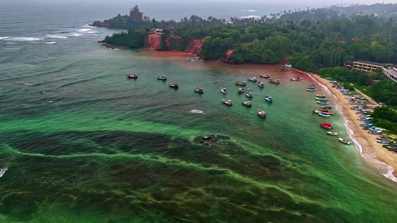 Vista aérea de um litoral curvo com vegetação exuberante e águas oceânicas azul-turquesa, barcos de pesca ancorados perto da praia arenosa de Weligama