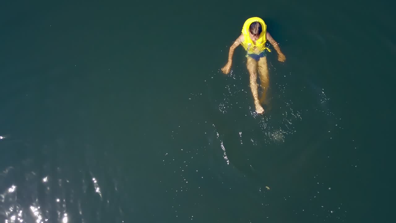Active kid in life yellow waistcoat swimming alone in the clear water in bright summer day. Portrait of cute funny boy bathing on his back in water swinging legs. Motion camera bottom up