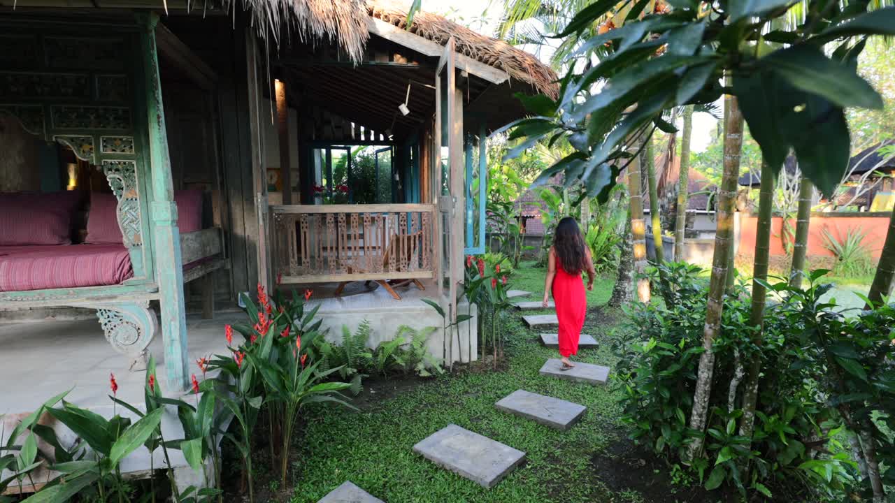 joven asiática vestida de rojo caminando por un sendero a través de un jardín tropical en una villa tradicional bali