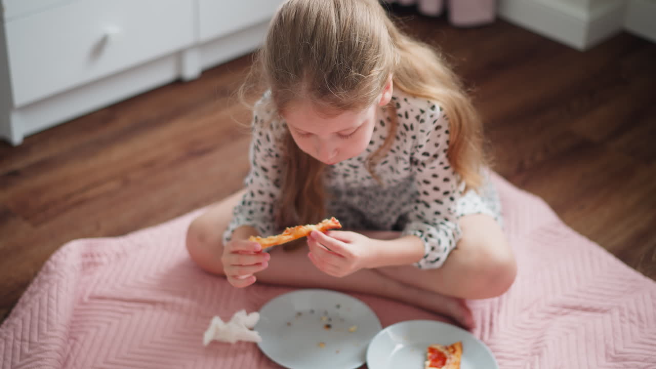 Kid sitting cross-legged on fluffy pink bedspread eating cheesy snack from plate, indoor setting with cozy wooden floor, spotted outfit, natural lighting