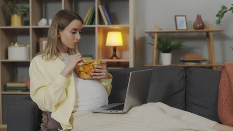 Pregnant woman watching laptop and eating chips