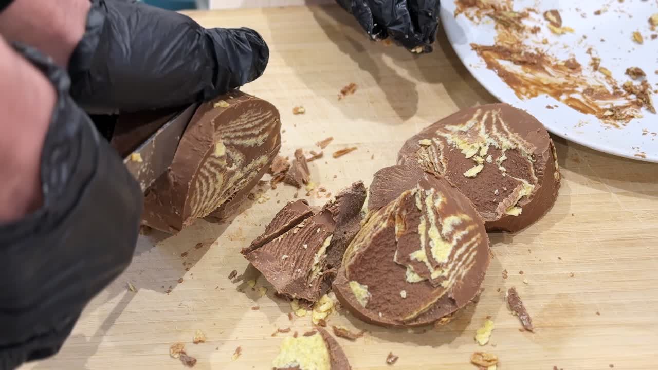 Primo piano di mani guantate che tagliano un dessert al cioccolato a strati su un tagliere di legno, con un interno cremoso, briciole e un piatto disordinato, sotto una luce soffusa da cucina interna.