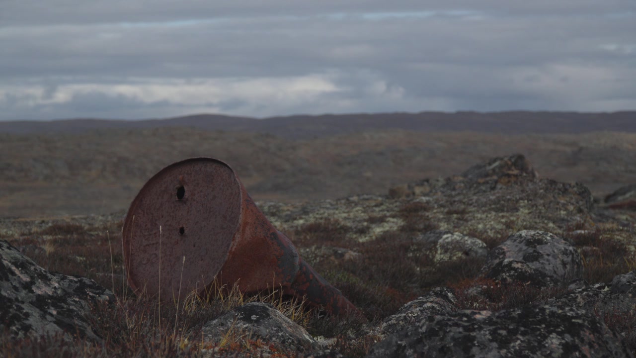 Ground level telephoto view focusing on weathered rusted steel drum sitting on tundra vegetation and granite rock
