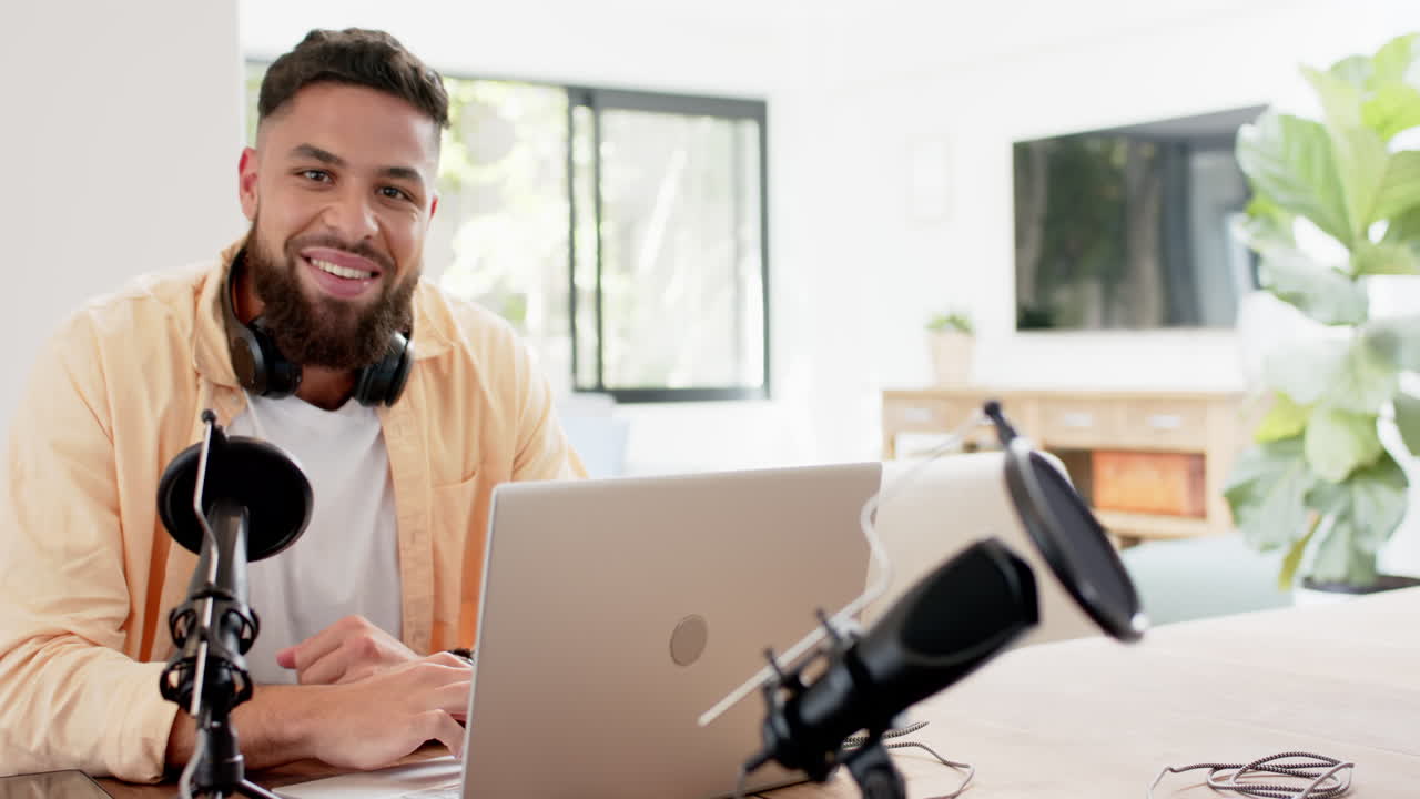 Smiling man using laptop and microphone for podcasting in home office