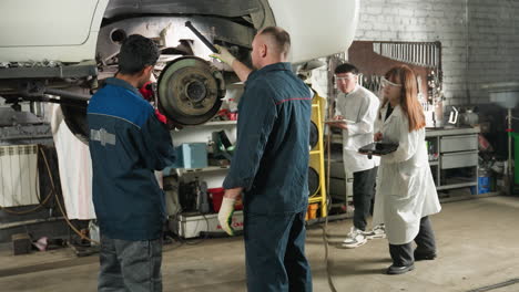 mechanic working on car repair while lab technicians with tablet observe and take notes, mechanical workshop setting with tools, lifted vehicle, collaboration, and technical inspection in progress