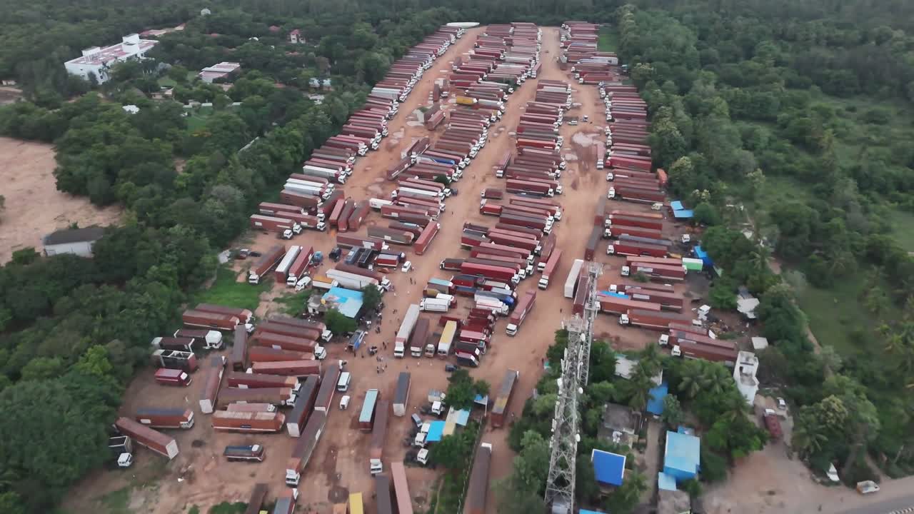 High-angle view of a huge truck parking lot carved from trees, adjacent to a major road network. Illustrates freight overflow and logistics management