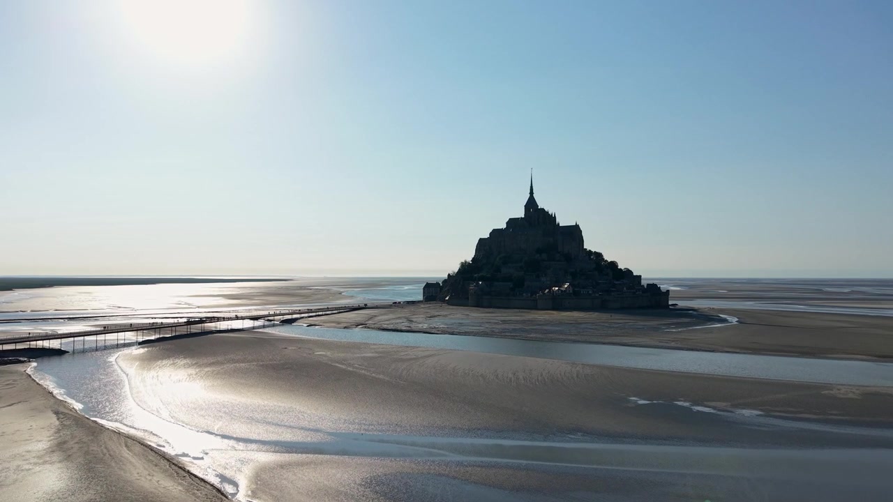 Vista aérea de drone do Mont Saint-Michel silhuetado contra o céu matinal, rodeado por vastas planícies de maré na maré baixa