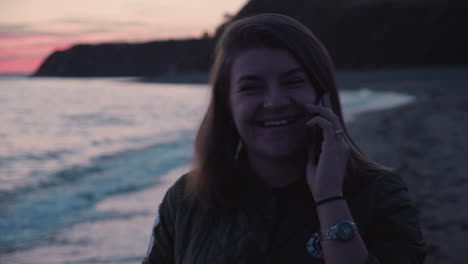 Una-Chica-Recibe-Una-Llamada-Telefónica-Y-Habla-Por-Teléfono-Mientras-Camina-Por-La-Playa-Al-Atardecer-Con-Olas-Del-Mar-Rompiendo-En-La-Orilla-Al-Fondo