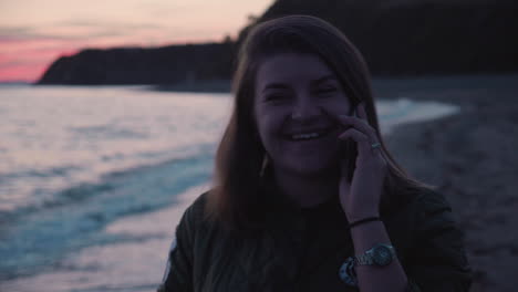 girl receiving a phone call and talking on the phone whie walking along the beach on the sunset with sea waves crushing on shore in the background