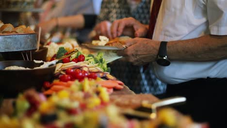 smooth shot of a line of people at a crowded event going through a food line and grabbing a variety of food including meats, cheeses, bread, fruits, and vegetables