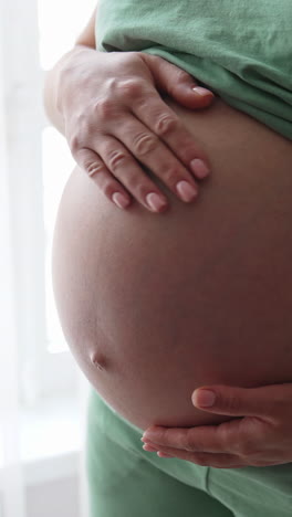 future mother standing by the large window. she's holding her big naked belly with one hand and stroking it with the other. close up. vertical video