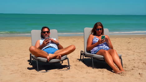 couple relaxing on beach chairs and using phones