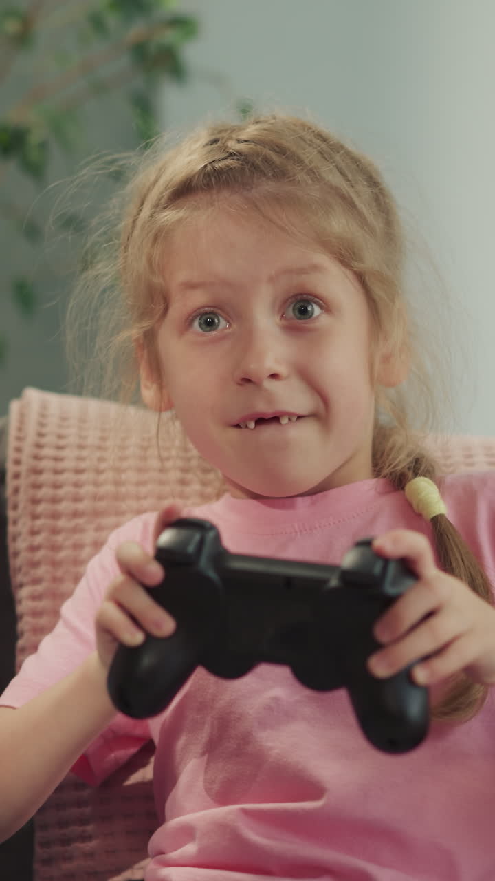 Toothless girl and blond boy play video game together sitting on sofa against pot-plant. Focused kids enjoy competing and having fun closeup