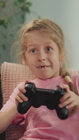 toothless girl and blond boy play video game together sitting on sofa against pot-plant. focused kids enjoy competing and having fun closeup